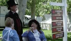 Three people dressed in old-fashioned, 19th-century clothing are gathered outdoors, smiling and talking. A man in a black suit and top hat stands next to two women seated on white chairs; one woman wears a blue dress and the other a blue and white outfit with a black hat adorned with flowers. Behind them, a wooden signpost points to different attractions, including "The Watering Hole," "Hart Hall," "SCV History Center," "Gold Rush Games," and "The Horseshoe Honky Tonk." White tents and green trees fill the background, at the annual Cowboy Festival